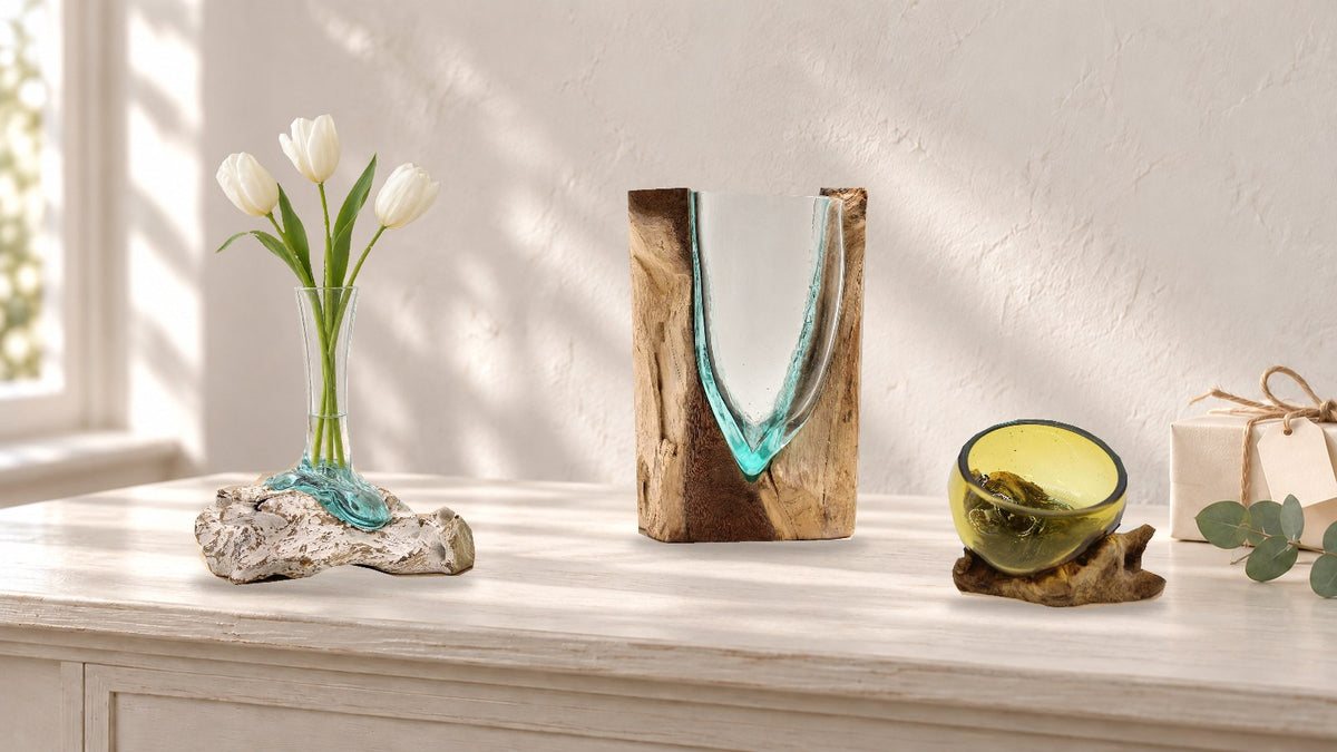 Molten glass vase on whitewashed wood holding three white tulips, a V-shaped glass vase in an upright block of wood and a mini amber glass bowl on wood, on a pale console
