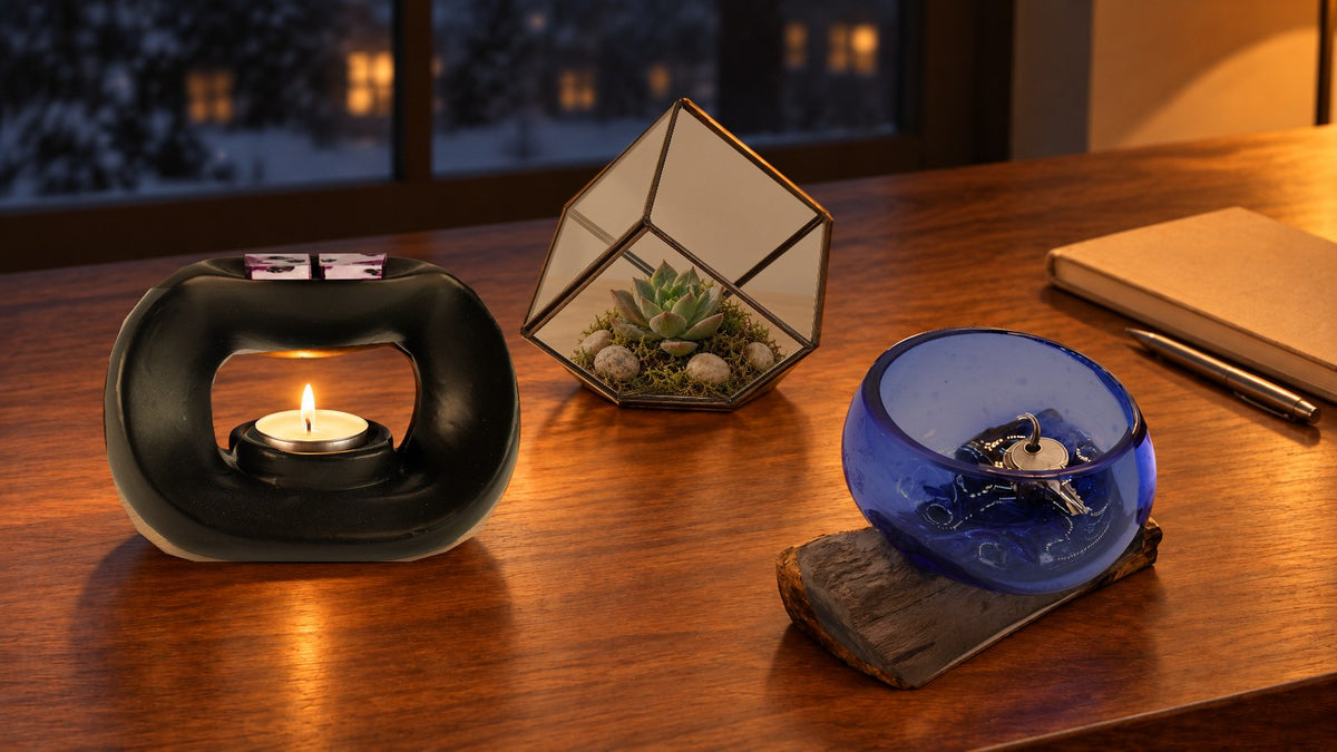 Matt black pebble wax burner with a lit tealight and wax melts, a cube terrarium with a succulent and a blue glass bowl on wood holding keys on a study desk at night