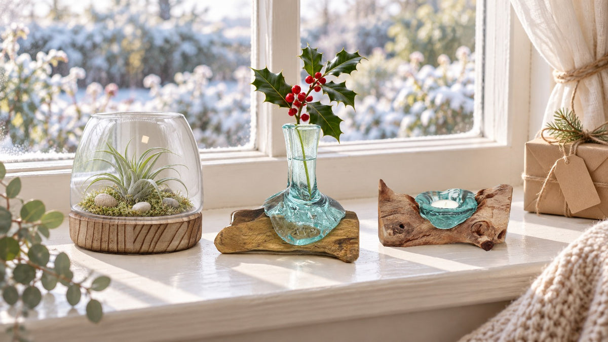 Small glass flower vase on wood holding a sprig of holly between a terrarium with an air plant on a tung wood base and a glass tealight holder on wood, on a cottage windowsill