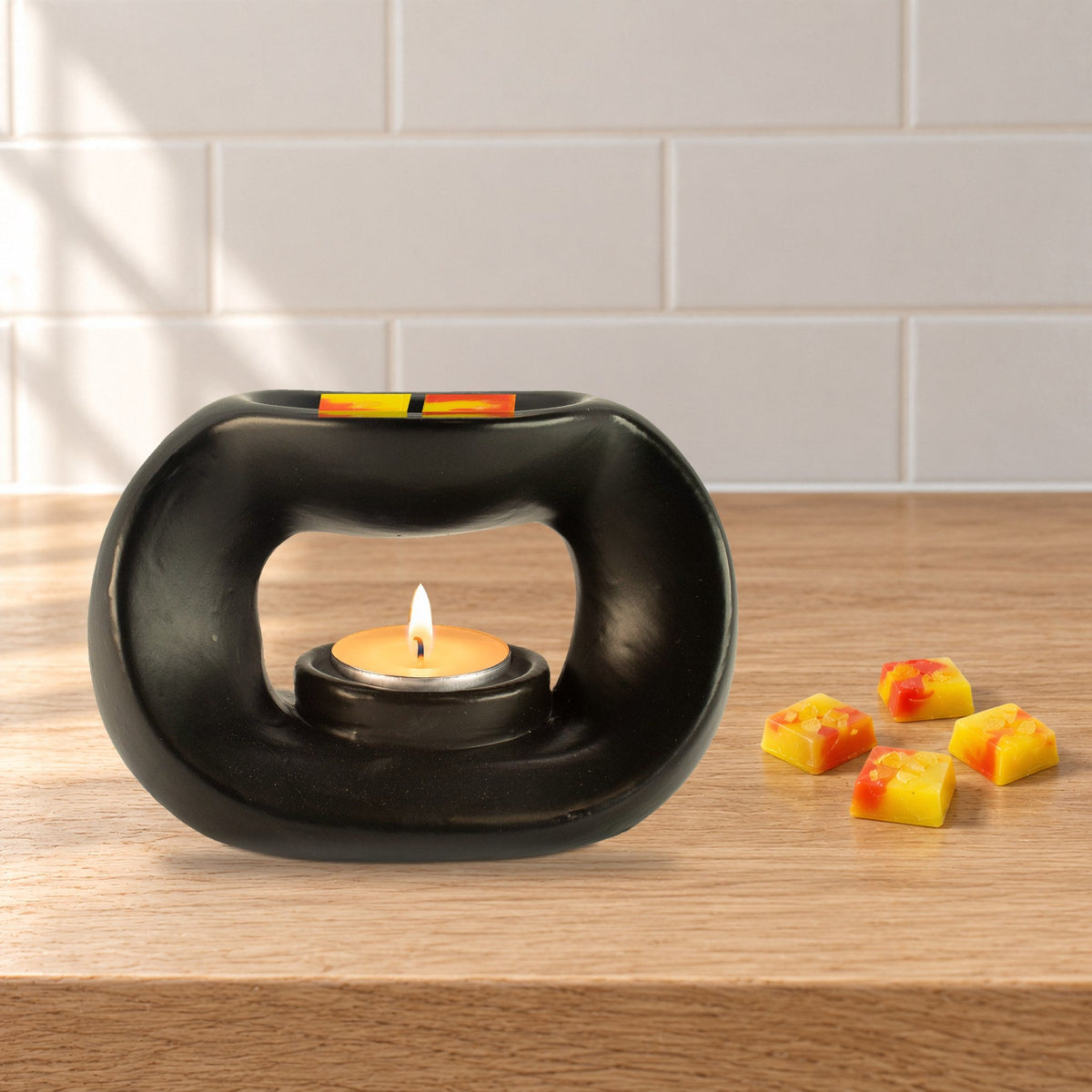 Matt black pebble wax burner with a lit tealight and Golden Glow yellow and orange wax melt squares, on a pale oak kitchen worktop