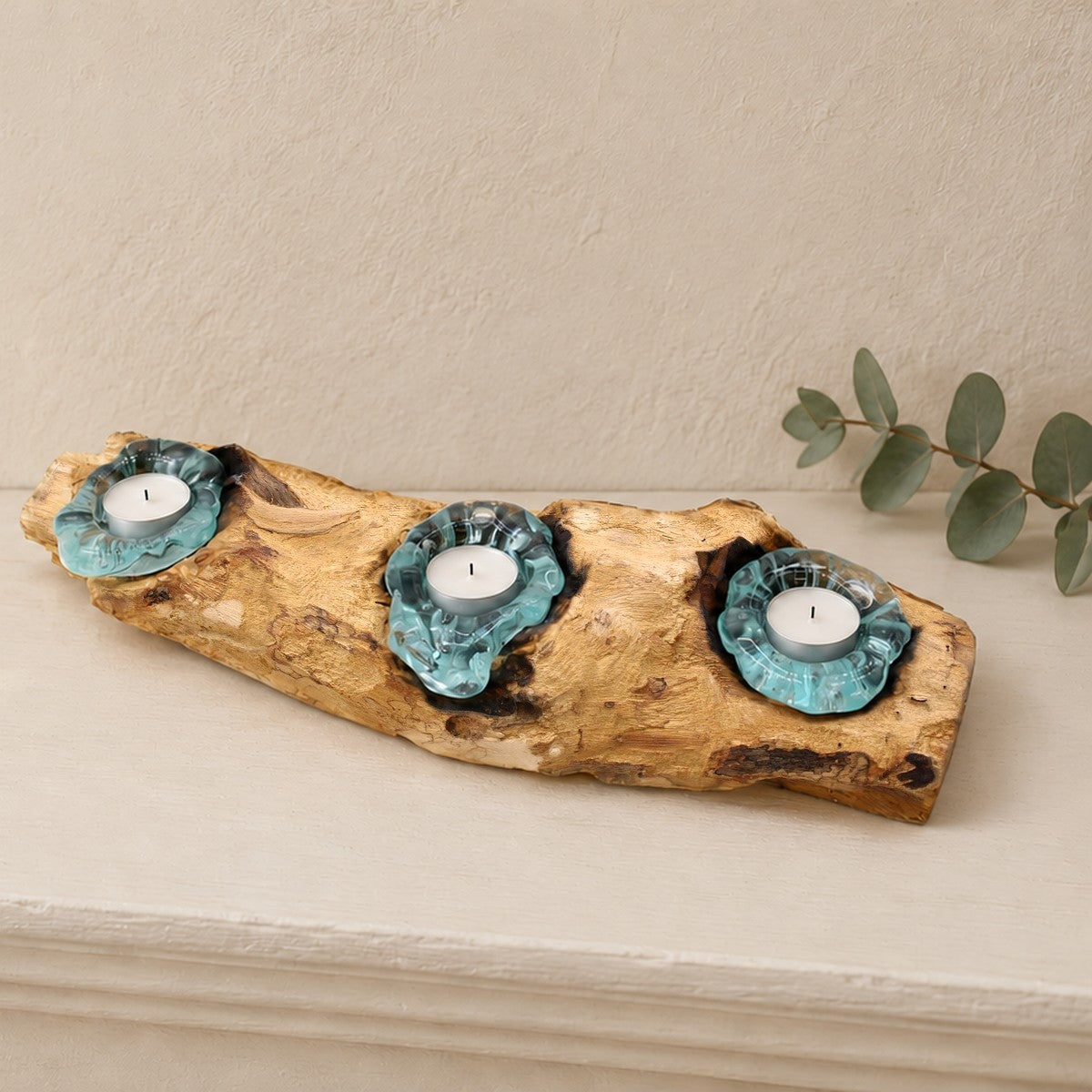Triple molten glass tealight holder on gamal wood with three unlit tealights, on a mantelpiece beside eucalyptus