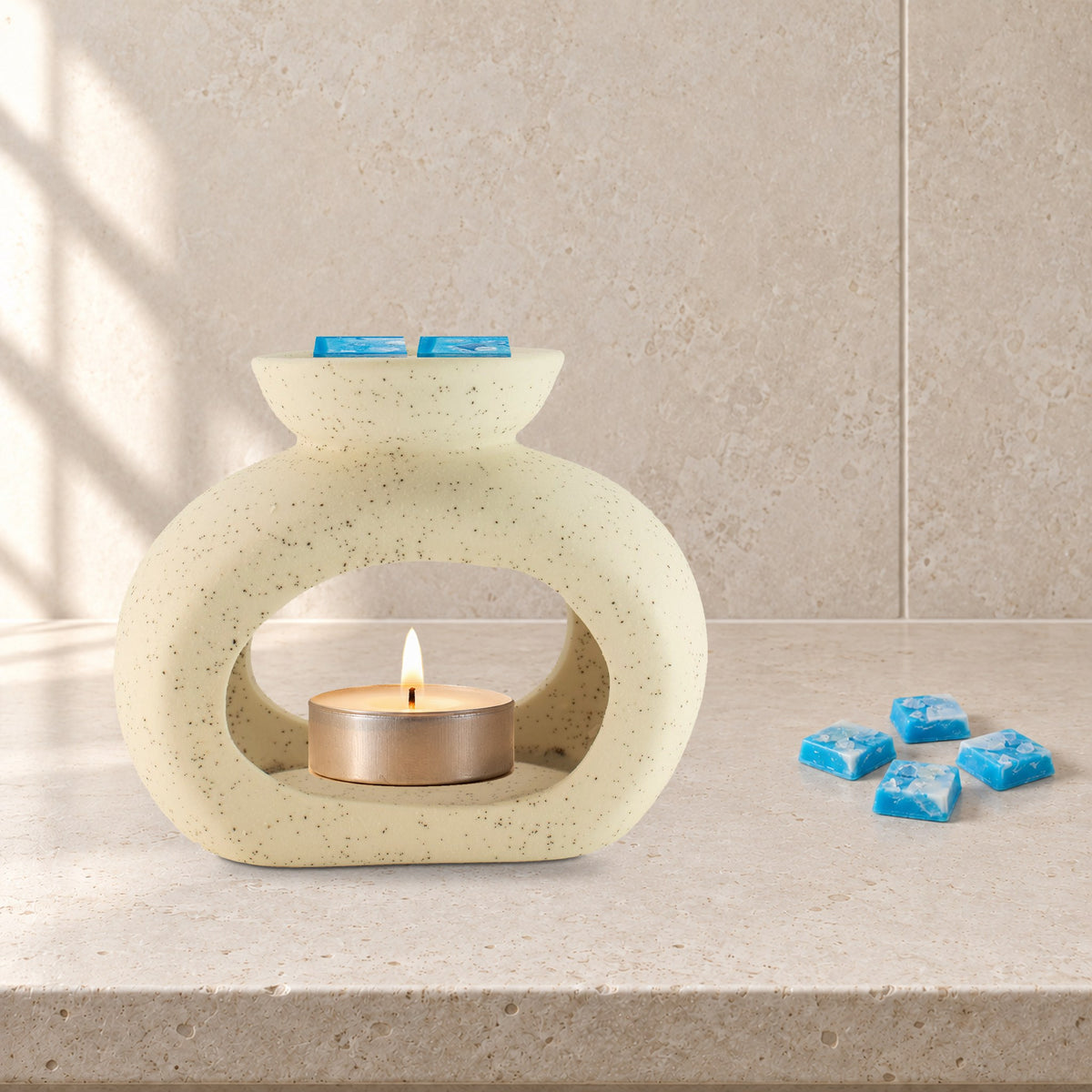 Speckled pebble wax burner with a lit tealight and Aquamarine Wave blue wax melt squares, on a pale stone bathroom shelf