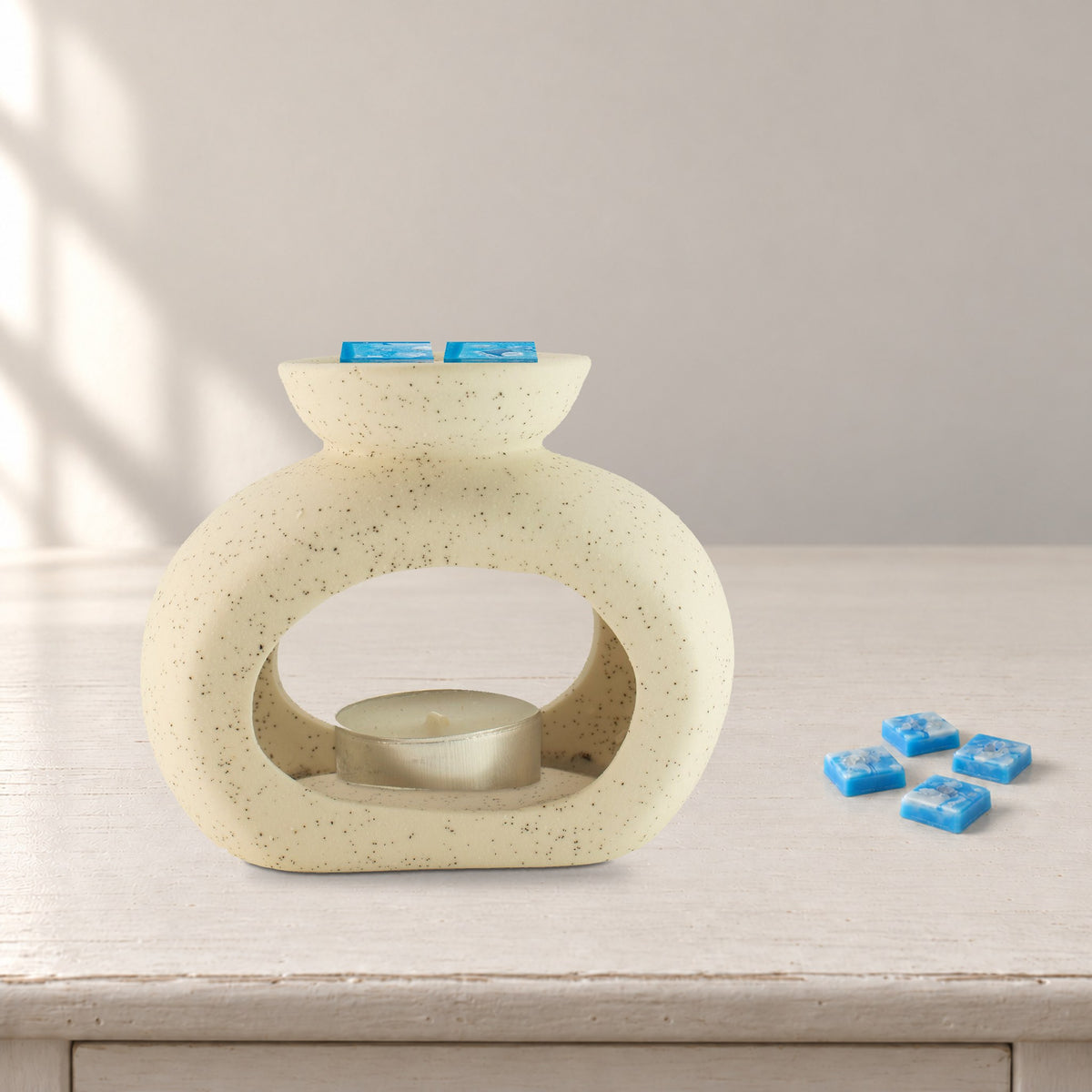 Speckled pebble wax burner with Aquamarine Wave blue wax melt squares and an unlit tealight, on a painted bedside table