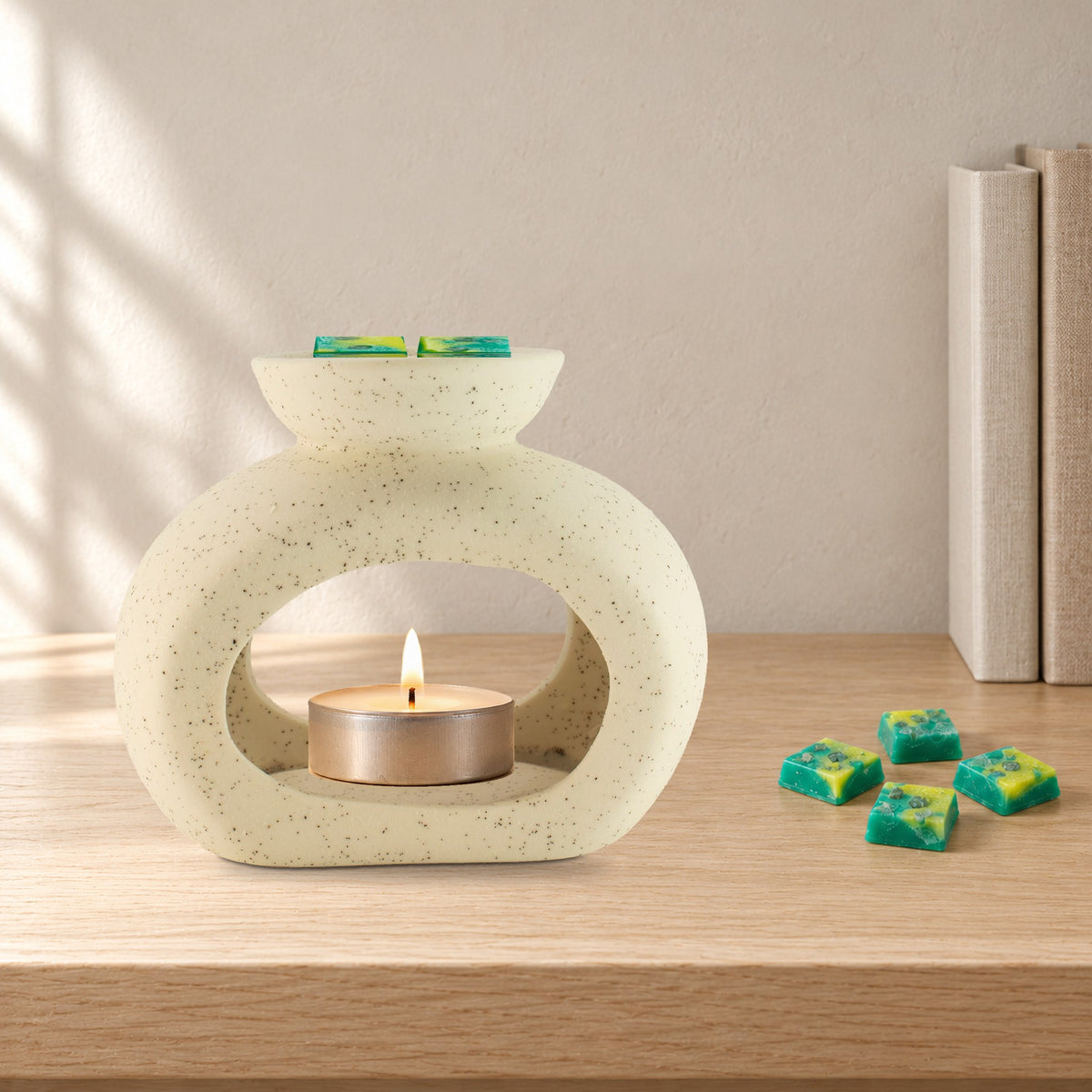 Speckled pebble wax burner with a lit tealight and Emerald Forest green and yellow wax melt squares, on a pale oak bookshelf