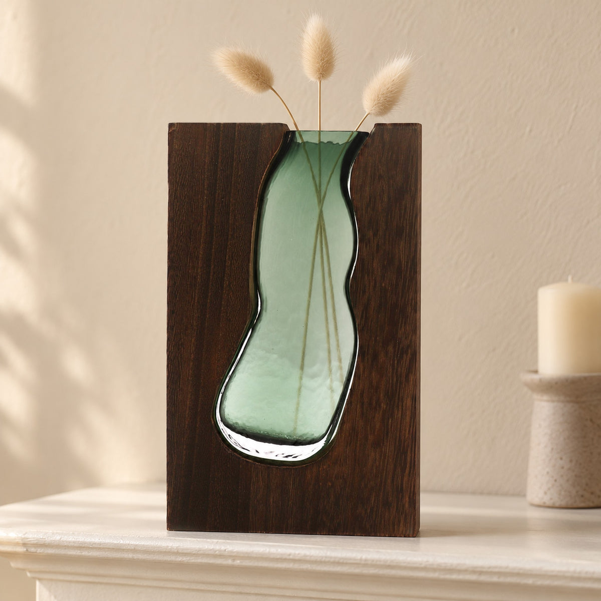 Large green glass vase terrarium set into dark tung wood, holding dried bunny-tail grass on a mantelpiece