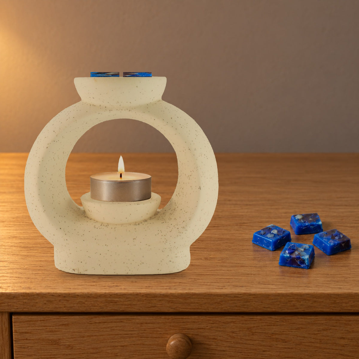 Large speckled pebble wax burner with a lit tealight and Lapis Night royal blue wax melt squares, on an oak bedside table
