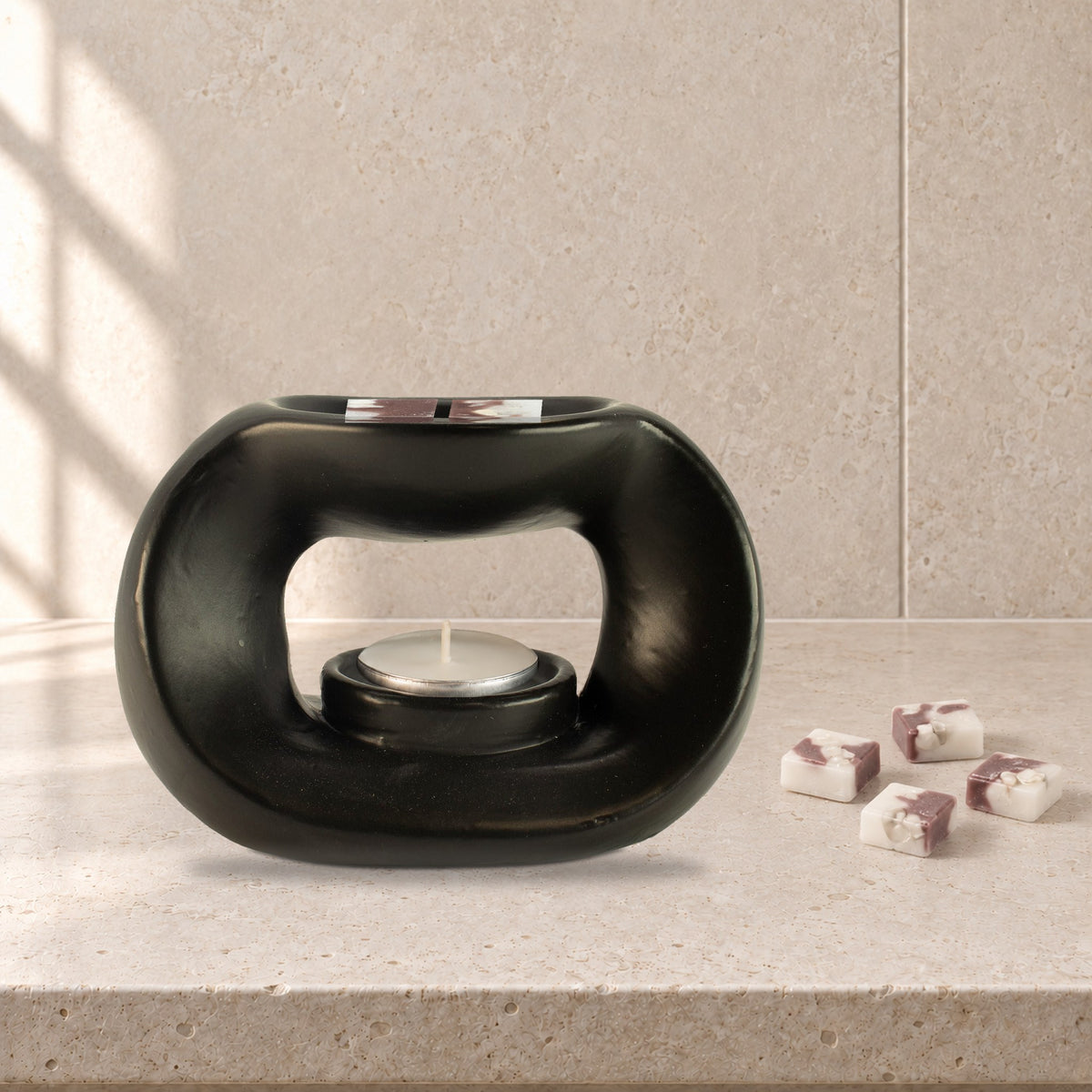 Matt black pebble wax burner with Moonstone Glow white and mauve wax melt squares and an unlit tealight, on a pale stone bathroom shelf