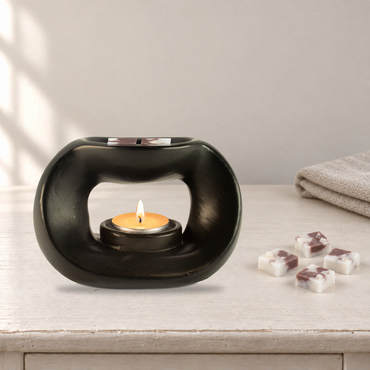 Matt black pebble wax burner with a lit tealight and Moonstone Glow white and mauve wax melt squares, on a painted dressing table