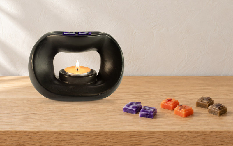 Matt black pebble wax burner with a lit tealight, two purple wax melt squares in its dish and purple, orange and brown wax melt squares beside it on an oak side table