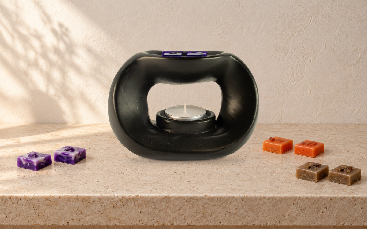 Matt black pebble wax burner with an unlit tealight and purple, orange and brown wax melt squares along a pale stone shelf